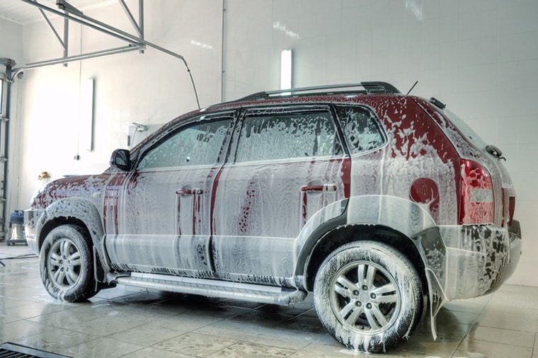 A red and silver SUV is covered in white soap suds while being washed in an indoor car wash facility. The car is on a wet tiled floor, and there is a partially open garage door visible on the left side. Bright lights illuminate the clean white walls.