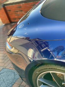 Close-up of the rear-left side of a shiny blue sports car, reflecting its surroundings like a brick wall and trees in its polished surface. The image highlights the car's sleek design, curving lines, and part of a rear tire labeled "305/30 ZR21.