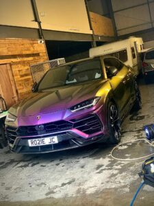 A purple Lamborghini Urus is parked in a large garage with wood paneling, white camper van, and other vehicles visible in the background. The license plate reads "RO21E JC". The car has a gradient color scheme and sporty design.