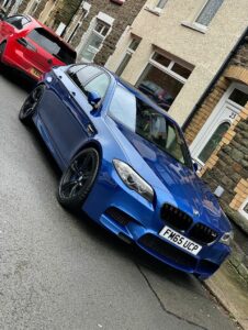 A blue BMW sedan, parked on a narrow residential street in front of terraced houses. Another red car is parked ahead of the BMW. Both cars have UK license plates, and the street is slightly wet.