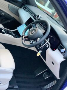 The image shows the interior of a modern car, featuring a sleek steering wheel with a key inserted, a digital dashboard, and a touchscreen infotainment system. The seats are upholstered in light-colored leather, and the floor has black carpeting.