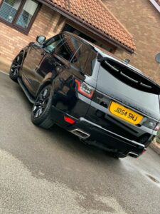 A black Range Rover is parked on a driveway in front of a brick house with a tiled roof. The vehicle has a yellow license plate with the registration "J054 SHU" and tinted windows. The ground appears wet as if it has recently rained.