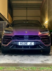 A sleek, purple Lamborghini SUV is parked inside a dimly-lit garage. The front view reveals the vehicle's bold design, with its headlights on, and a UK license plate reading "RO21E JC" clearly visible.