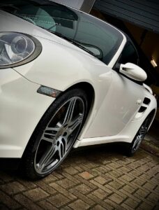 A white sports car is parked on a brick driveway. The image is taken from a low angle, showcasing the car's sleek side profile and front wheel. The garage door in the background is slightly open, revealing a glimpse of the interior.