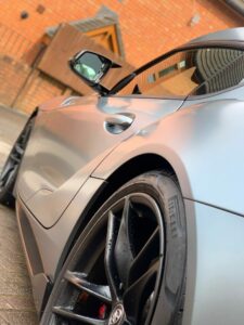 Close-up angle of a sleek silver sports car parked in front of a brick wall and wooden structure. The focus is on the car's rear wheel and side mirror, showcasing its aerodynamic design, sharp lines, and black Pirelli tires.