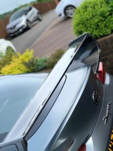 Close-up of the rear of a silver Toyota Supra focusing on its large carbon fiber spoiler. The car is parked in a driveway with other vehicles and greenery visible in the background. The image captures the sleek and sporty aesthetic of the vehicle.