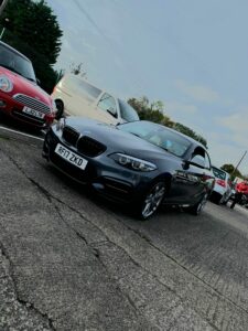 A dark grey BMW with the license plate RF17 ZKD is parked on a concrete surface. Surrounding the BMW are a red Mini Cooper, a white van, and other vehicles in a parking lot under an overcast sky. Trees and a chain-link fence are visible in the background.