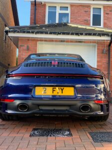 A blue Porsche 911 with a yellow license plate is parked on a brick driveway in front of a modern red-brick house with white-framed windows. The house has string lights hanging from the roof and a closed garage door.