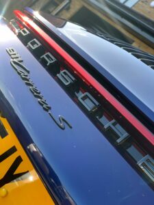 Close-up of the rear section of a dark-colored Porsche 911 Carrera S, showing the Porsche logo, model name, and part of the brake light. The car is parked near a building, and part of a yellow license plate is visible at the bottom left of the image.