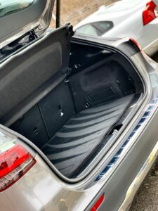 An open car trunk showing an empty and clean cargo space. The trunk is part of a silver car with a portion of a white car visible in the background. The interior of the trunk has a black lining and appears spacious.