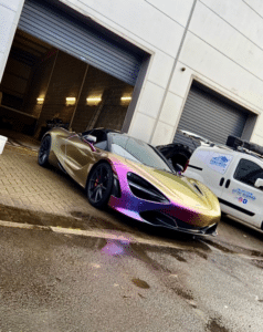 A sleek, metallic-colored sports car with a gradient paint job ranging from gold to purple is parked in front of a garage. The car has its convertible top down and is positioned next to a white van. The ground around is wet, and there are two large garage doors behind.