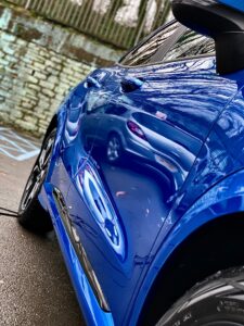 Close-up of the side of a shiny blue car, showing the reflection of a white car and tree branches on its surface. The setting appears to be an outdoor parking area with a wet ground, a stone wall, and a metal fence in the background.