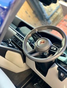 Interior view of a car showing the steering wheel with the Porsche logo. The dashboard features a modern digital display, control buttons, and air vents. The interior has a sleek and luxurious design with a mix of light and dark materials.