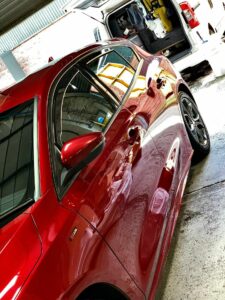 A red car is parked under a shelter, reflecting its surroundings. A partially open van with equipment and supplies inside is visible in the background. The scene appears to be a garage or vehicle maintenance area.