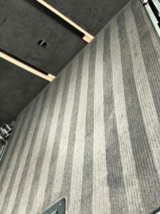 A view of a carpeted floor with a subtle striped pattern in shades of gray. The image is taken at an angle, emphasizing the texture and design of the carpet. Part of a dark wall or piece of furniture is visible in the background.