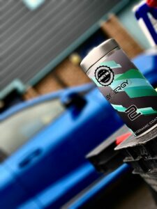 Close-up of a bottle of car care product labeled "Synergy 2" on a surface with a blurred blue car in the background. The bottle features a green and black design with a silver cap and the words "ceramic coating" visible at the bottom.