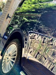 Close-up of the side of a shiny black car parked near a wooden structure. The car's surface reflects leafy green trees and a cobblestone path. The focus is on the polished surface and the detailed pattern of the tires and rims.