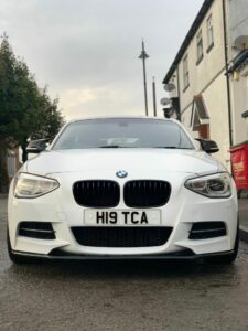 A white BMW car is parked on a street near houses. The car's license plate reads "H19 TCA." The street is slightly wet, suggesting recent rain. The sky is overcast, and there are trees and a red bin visible in the background. The car is centered and facing forward.