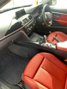 Interior view of a car featuring a modern dashboard with a digital display, a center console with control buttons, and red leather seats. The view shows the driver's side, passenger seat, and dark floor mats. The car has a sleek, sporty design.