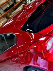 A shiny red car with a closed back door and a visible taillight is parked indoors. The car's surface reflects light, highlighting its glossy finish. Shelves and various items in the background indicate the car is in a garage or workshop.