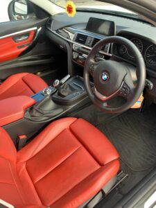 Interior view of a BMW car featuring a black and red leather seating. The dashboard includes a central digital display, surrounded by various controls. The steering wheel displays the BMW logo, and the gear shift is located between the front seats.