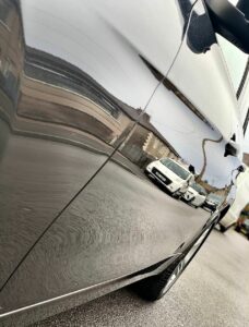 A close-up view of the side of a silver car, showing its well-maintained exterior, which reflects several cars parked along the street. The reflection is slightly distorted, but nearby buildings and the road are visible in the car's shiny surface.