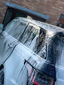 A dark-colored SUV is covered in soap suds, parked in front of a brick building. The vehicle appears to be in the process of being washed, with sunlight reflecting off its wet surface.