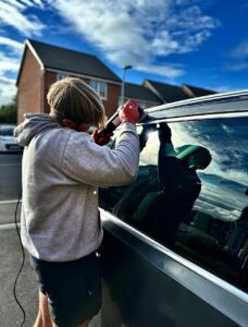 A person wearing a grey hoodie and red gloves is using a tool on the window of a car parked outside. The sky is partly cloudy, and houses can be seen in the background.