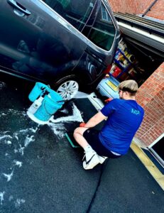 A person wearing a blue shirt and white shorts is washing a dark-colored car in a driveway. They are crouching down and holding a hose, spraying water on the car. A soapy bucket and other cleaning supplies are next to them. A garage with items inside is visible in the background.