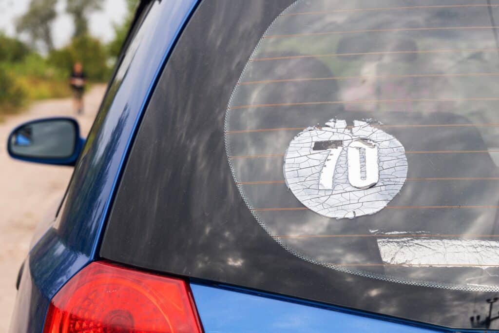 A close-up of the rear window of a blue car showcases a cracked sticker with the number "70" on it. A blurred figure is visible in the background on a rural road, contemplating sticker removal.