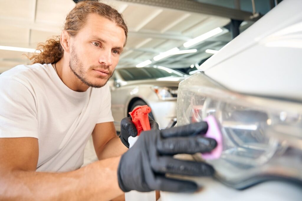 A person with long hair is wearing gloves and polishing a car's headlight in a garage. They are focused on their task, holding a pink cloth and a red spray bottle. The background shows other cars and bright ceiling lights.