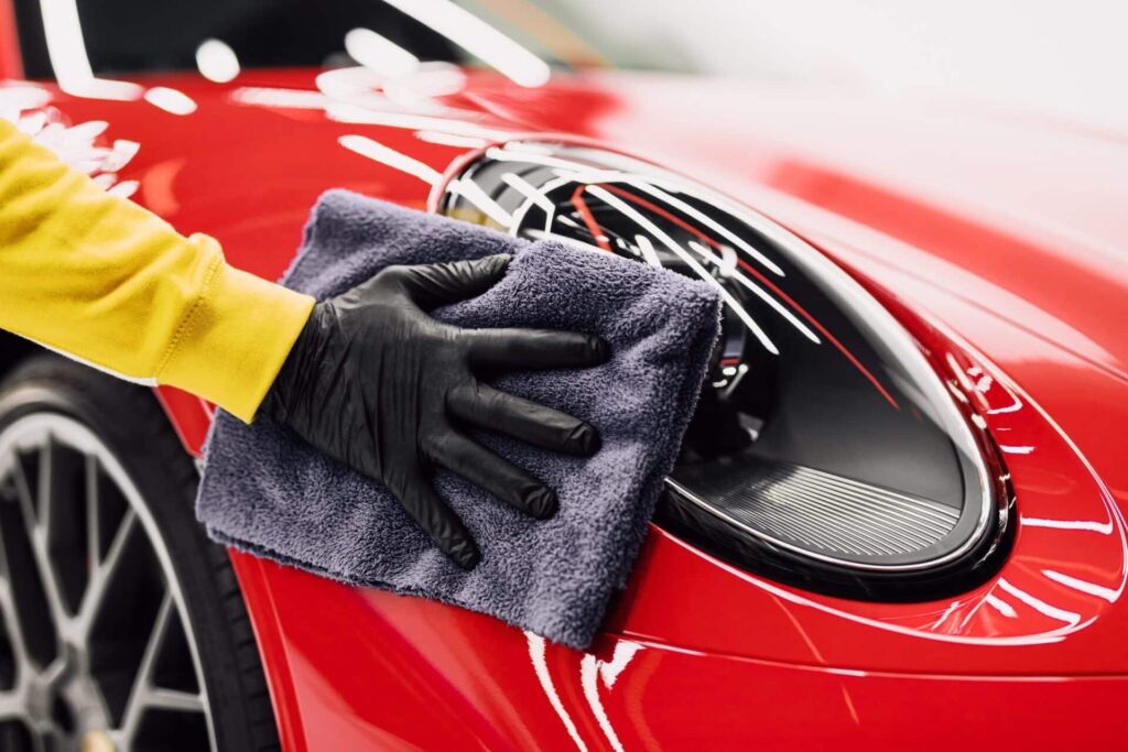 A person wearing a yellow sleeve and black glove polishes the headlight of a shiny red sports car with a gray cloth. The car's sleek design and circular headlight are prominently featured.